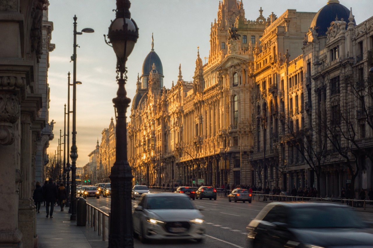 Pedestrians crossing a wide boulevard between stone facades in flat morning light